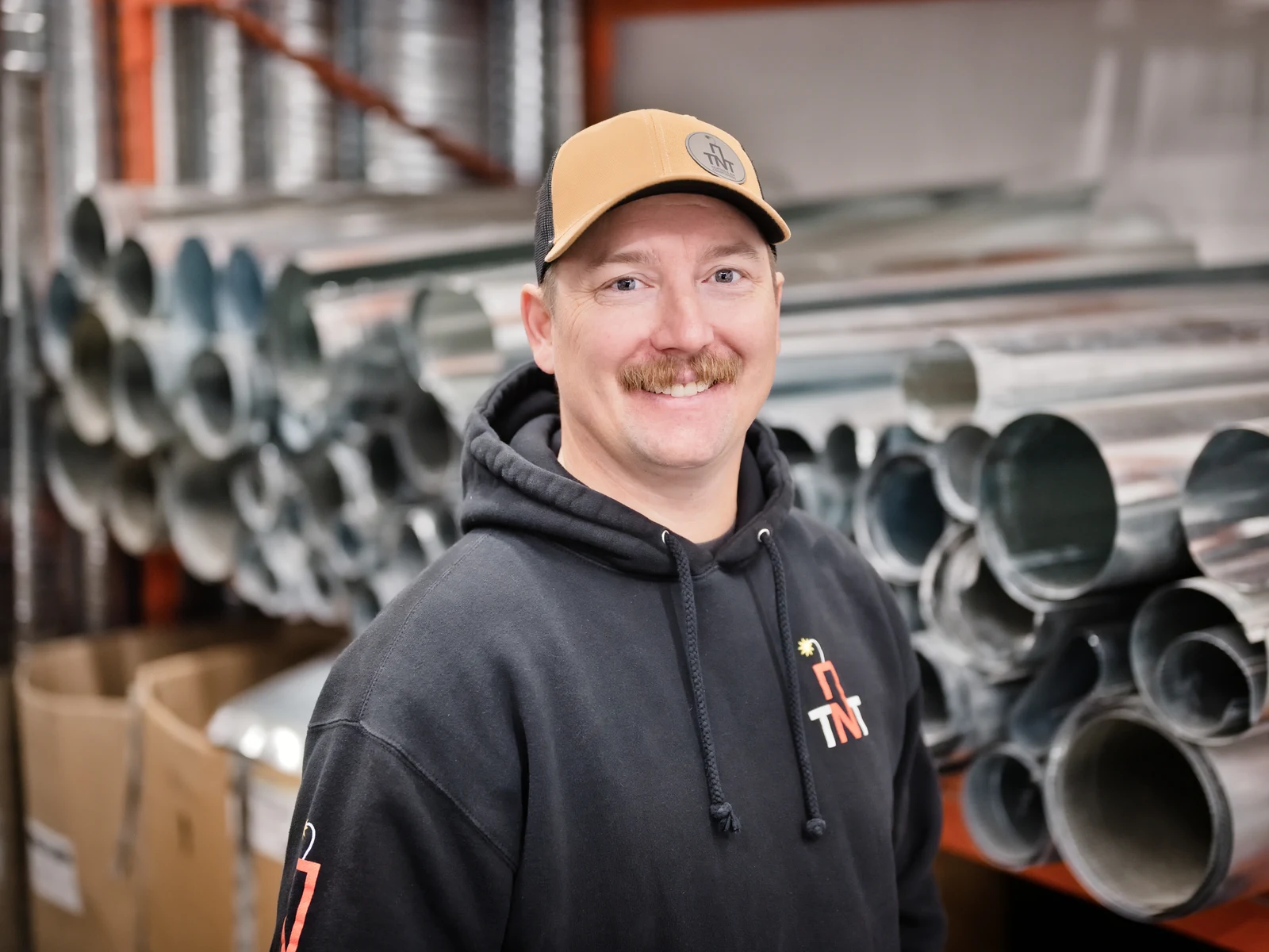 Furnace installation Calgary: one of the two TNT owners in the Calgary shop, in front of a stack of galvanised duct