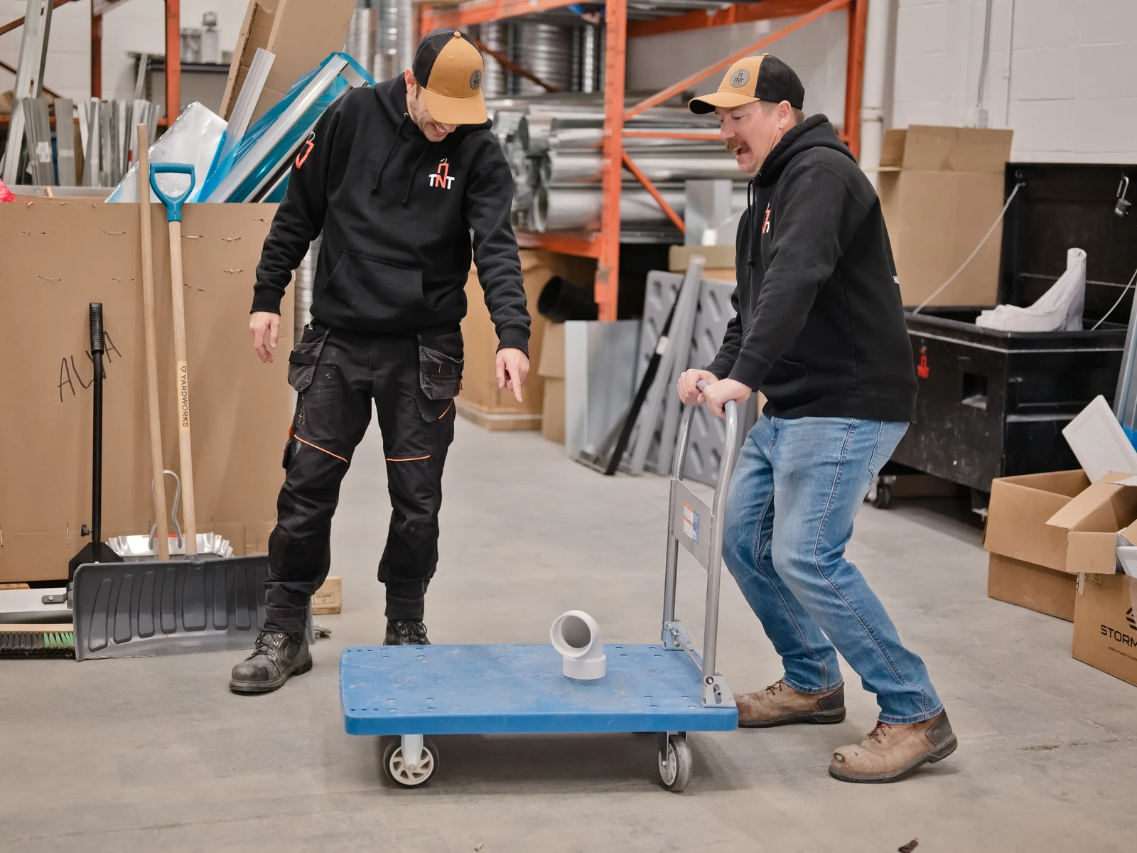 HVAC services Calgary: one TNT Heating owner riding a shop trolley while the other pushes it across their Calgary shop